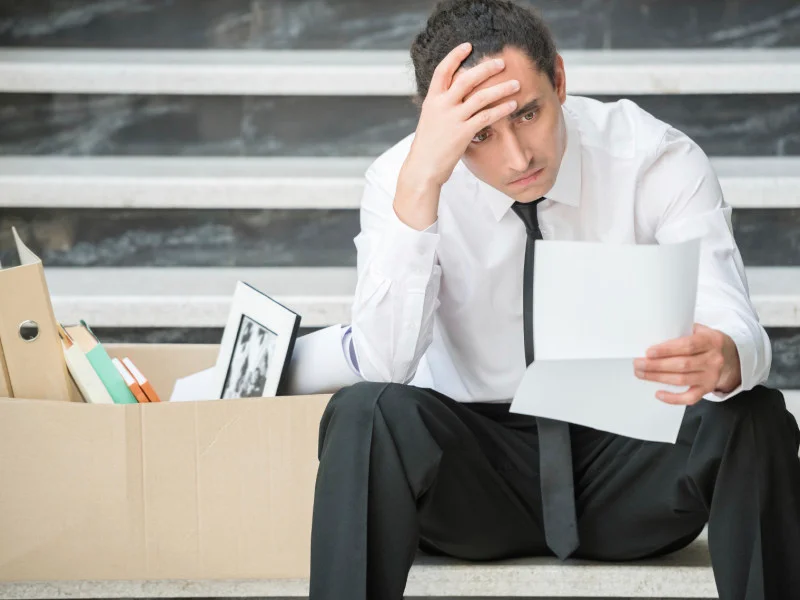 Business man holds his head as he sits slumped over looking at a piece of paper in his hand, belongings in a cardboard box next to him