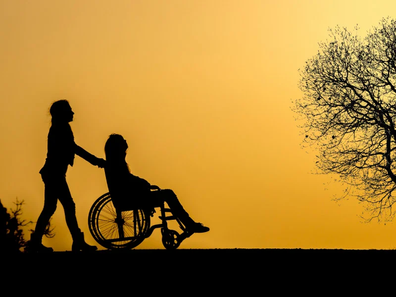 Silhouette of a person pushing another person in a wheelchair in front of a yellow sky