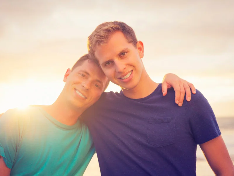 Two smiling men with their arms around each other in front of a sunset